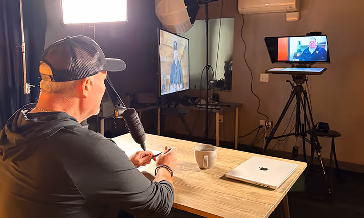 Man in a cap recording a podcast or video in a studio with professional lighting, a microphone, a laptop on the table, and two screens displaying another person.