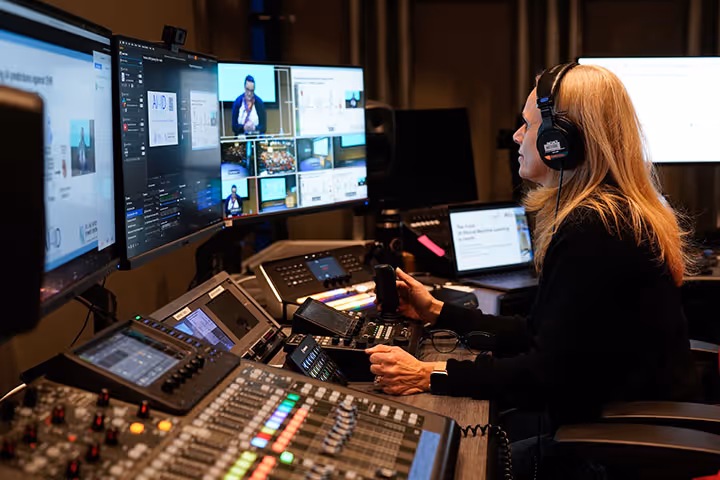 Woman with headphones operating a livestream control room with multiple monitors and audio mixing equipment.