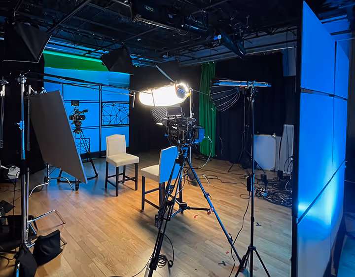 Indoor video production studio setup with cameras, lighting equipment, two beige chairs, and blue and green backdrop curtains.